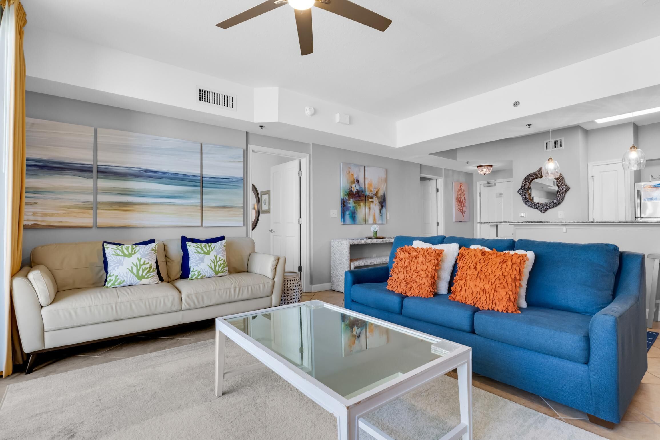 A modern living room with a beige sofa, a blue sofa with orange pillows, a glass coffee table, and abstract wall art.