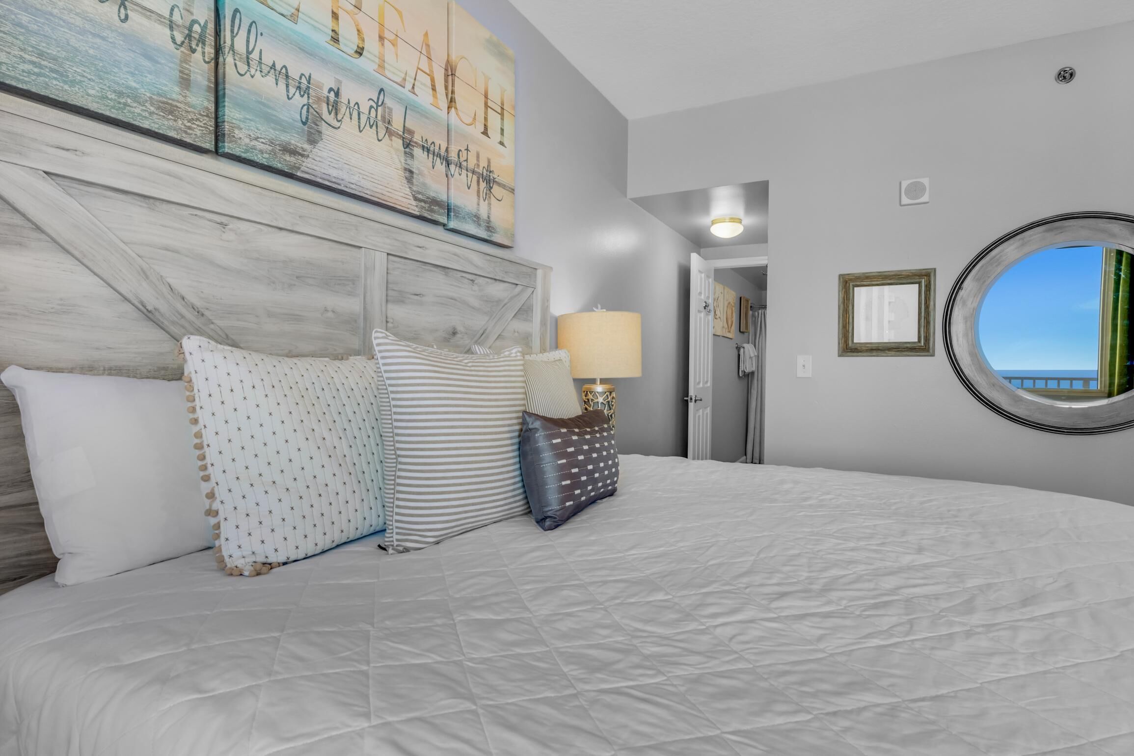 Een slaapkamer met een groot bed, een hoofdbord met een rustiek design, een ronde spiegel en uitzicht op zee door een raam.