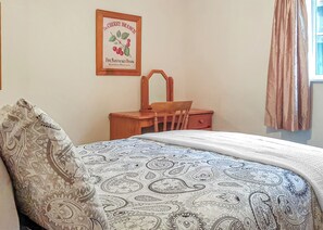 Un dormitorio con una colcha estampada, un escritorio de madera, una silla, un cuadro en la pared y una ventana con cortinas.