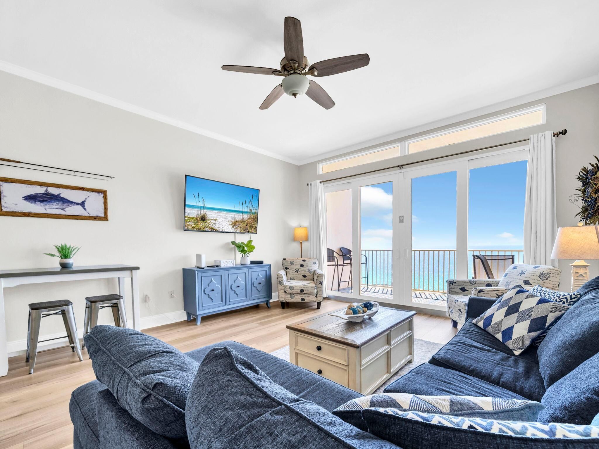 Amplia sala de estar con ventilador de techo, un ventanal con vistas a la playa, comedor con un mueble azul y zona de estar con cojines estampados.
