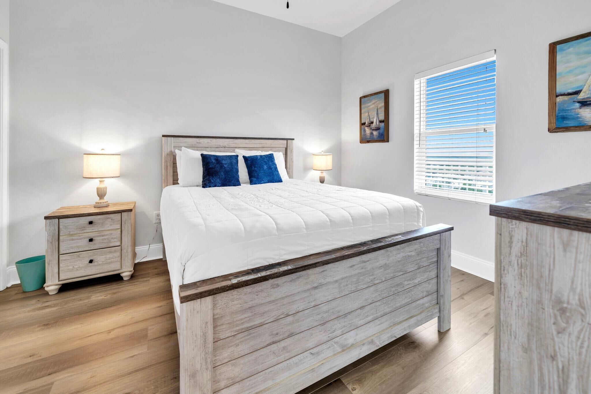 A bedroom with a bed, two bedside tables, a nightstand, a window with blinds, and a painting on the wall.