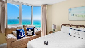 Ett hotellrum med ett stort fönster som vetter mot havet, en säng med vita sängkläder, en beige soffa med kuddar med marint tema och en tavla på väggen.