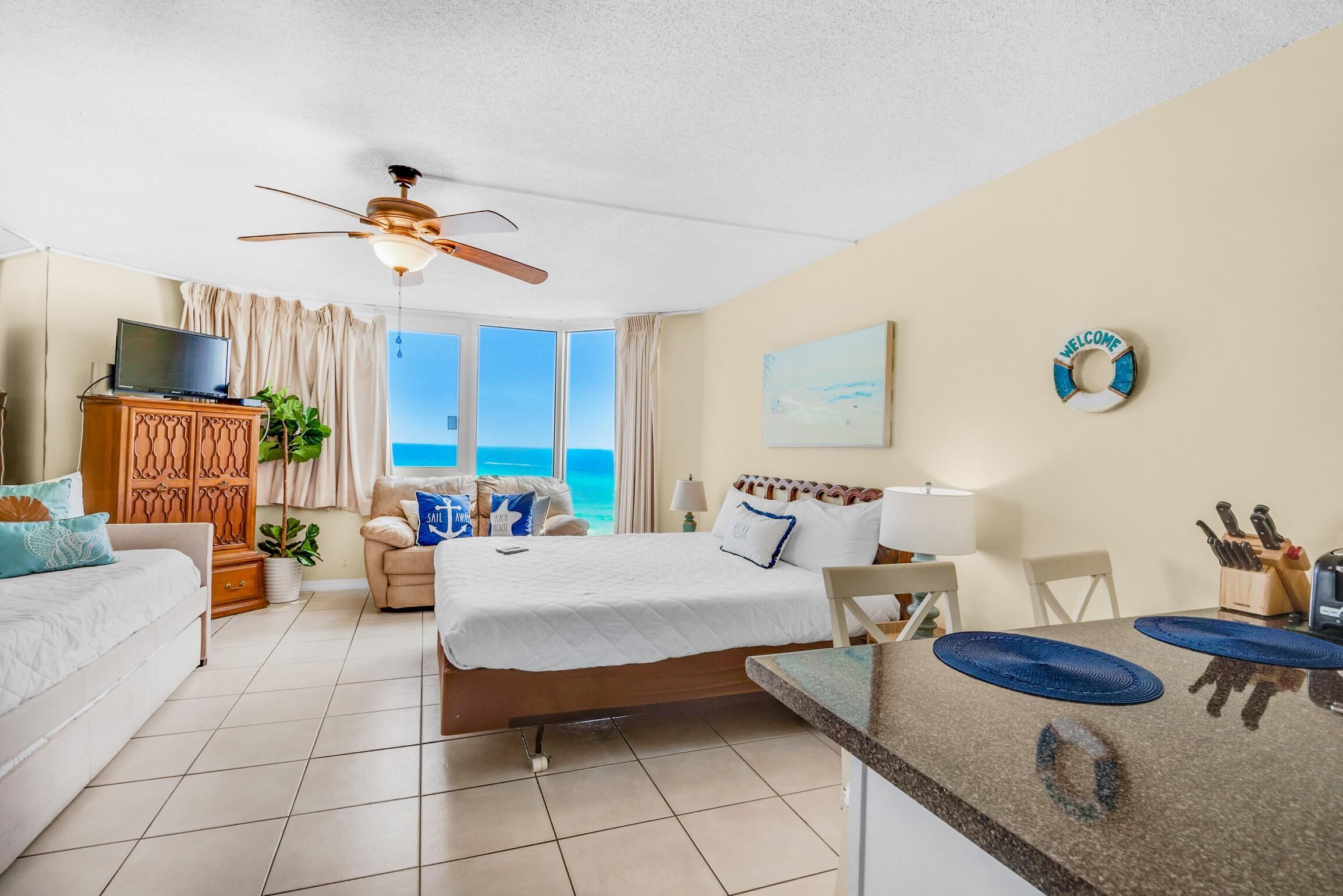 A hotel room with a bed, a desk, a TV, a ceiling fan, a view of the ocean, and a welcome sign.