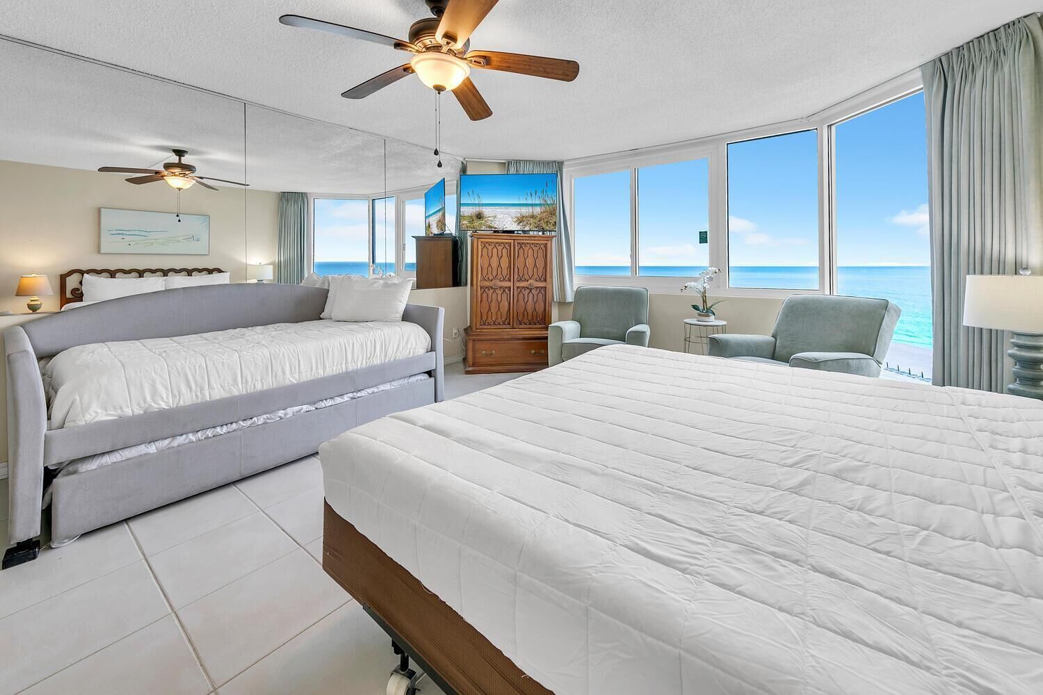Een hotelkamer met een groot bed, een slaapbank, een plafondventilator, een televisie, een houten kast en uitzicht op de oceaan.