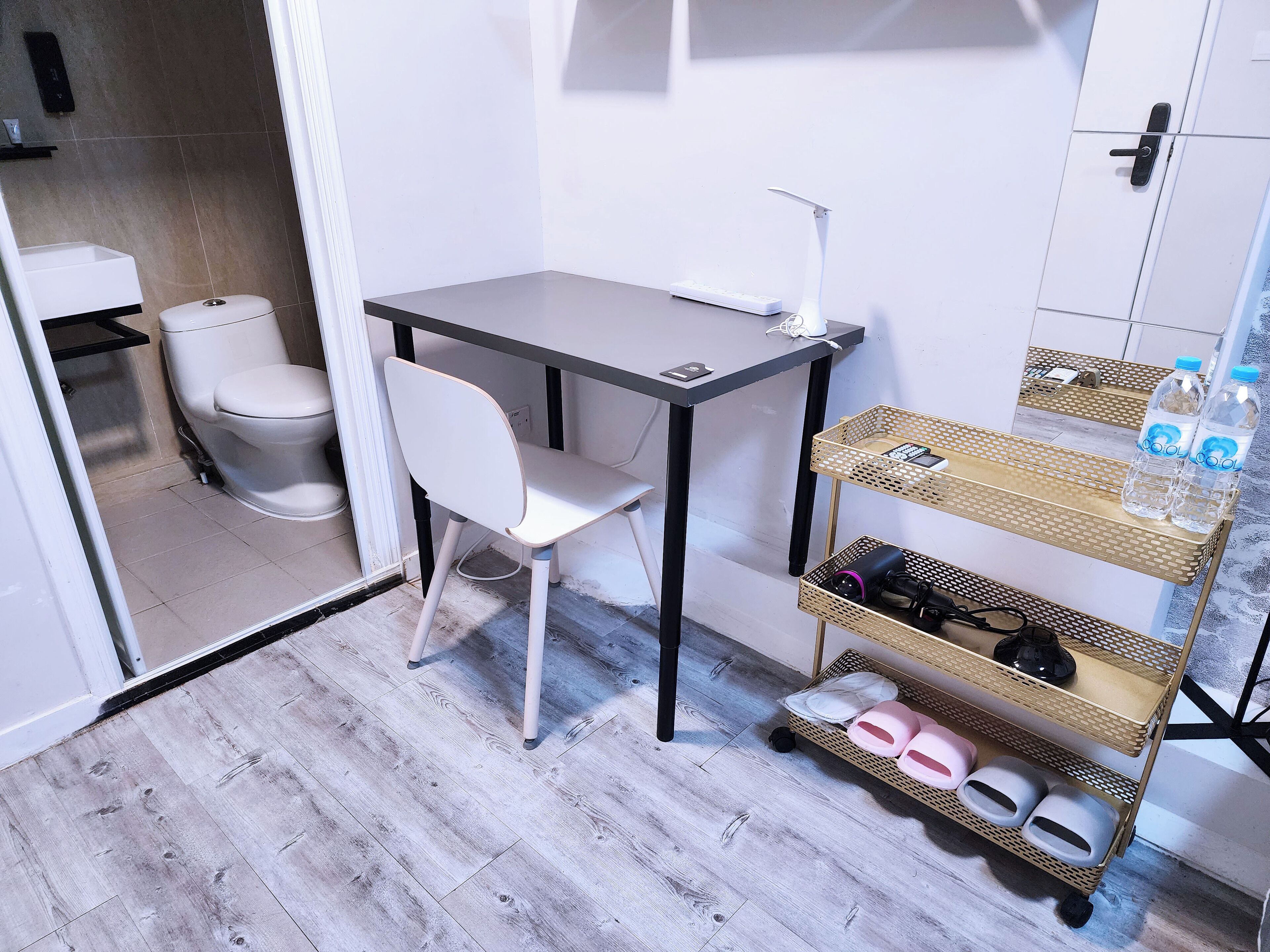 A small, modern bathroom with a white toilet, a white chair, a small table with a lamp, and a shelf with shoes and toiletries.