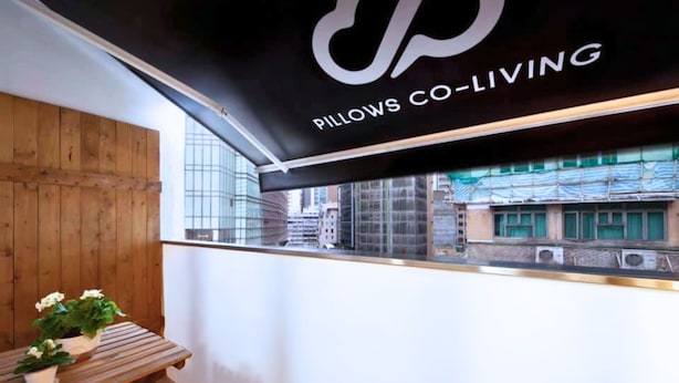 A modern interior with a black awning displaying "Pillows Co-Living" logo, a wooden table with potted plants, and a view of a cityscape through large windows. - Pillows Co-Living (Kowloon)
