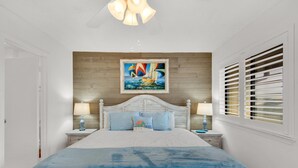 A bedroom with a large bed, wooden headboard, two bedside tables with lamps, a window with blinds, and a painting on the wall. - Sandprints A12 (Miramar Beach)