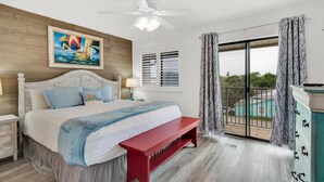 A bedroom with a bed, a red bench, a ceiling fan, a painting of sailboats, and a view of a pool through a sliding glass door. - Sandprints A12 (Miramar Beach)