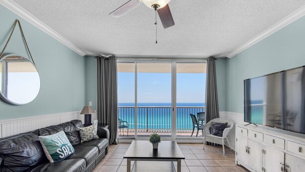 A living room with a view of the ocean, a ceiling fan, a TV, and a sofa with a pillow. - Majestic Beach Towers 1-1302 (Panama City Beach)