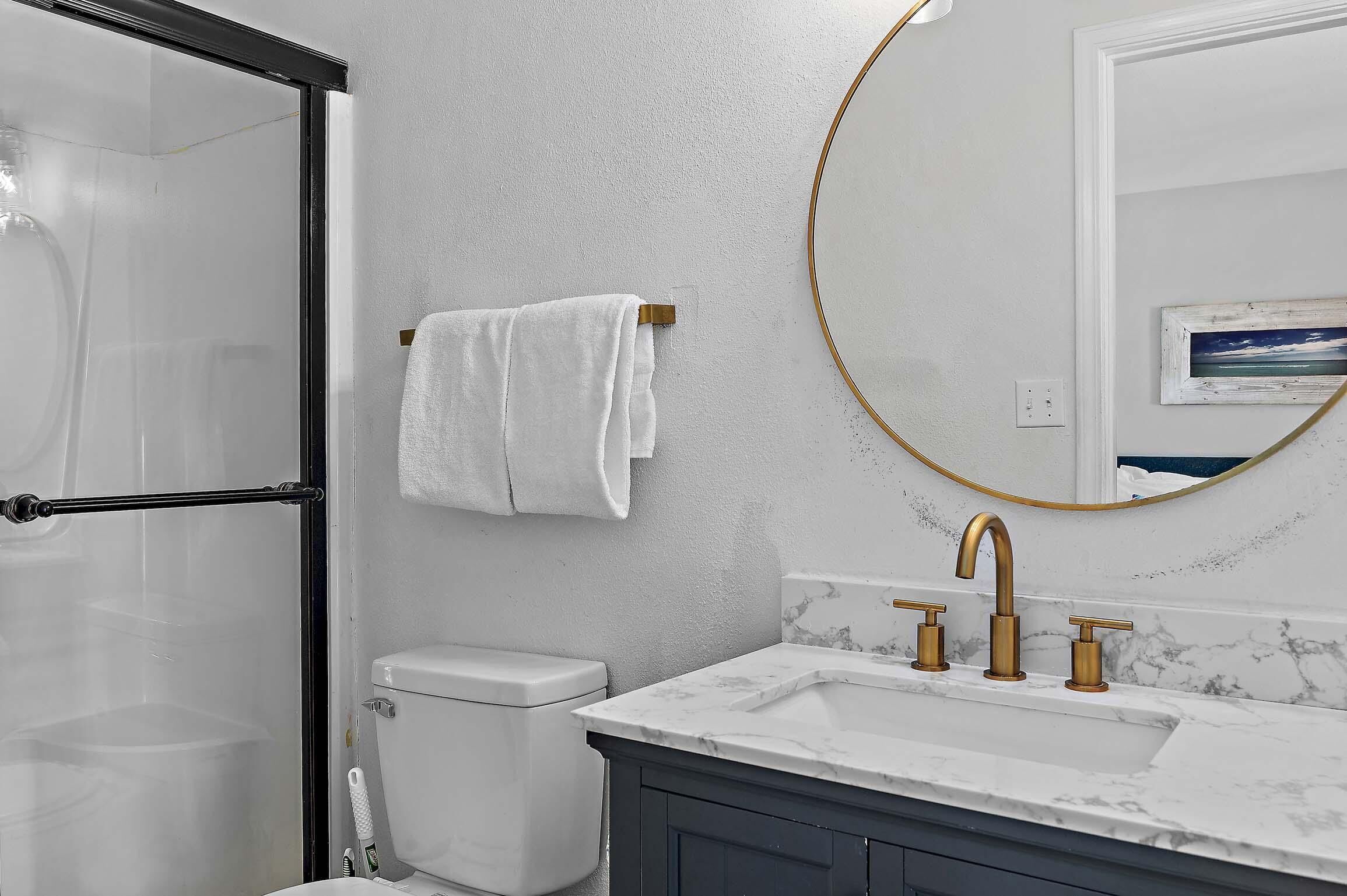 A bathroom with a white toilet, a shower with a glass door, a white towel hanging on a rack, a marble sink with gold faucets, and a round mirror with a gold frame.