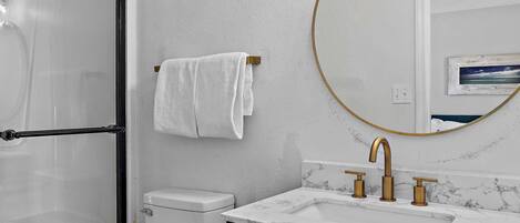 A bathroom with a white toilet, a shower with a glass door, a white towel hanging on a rack, a marble sink with gold faucets, and a round mirror with a gold frame.