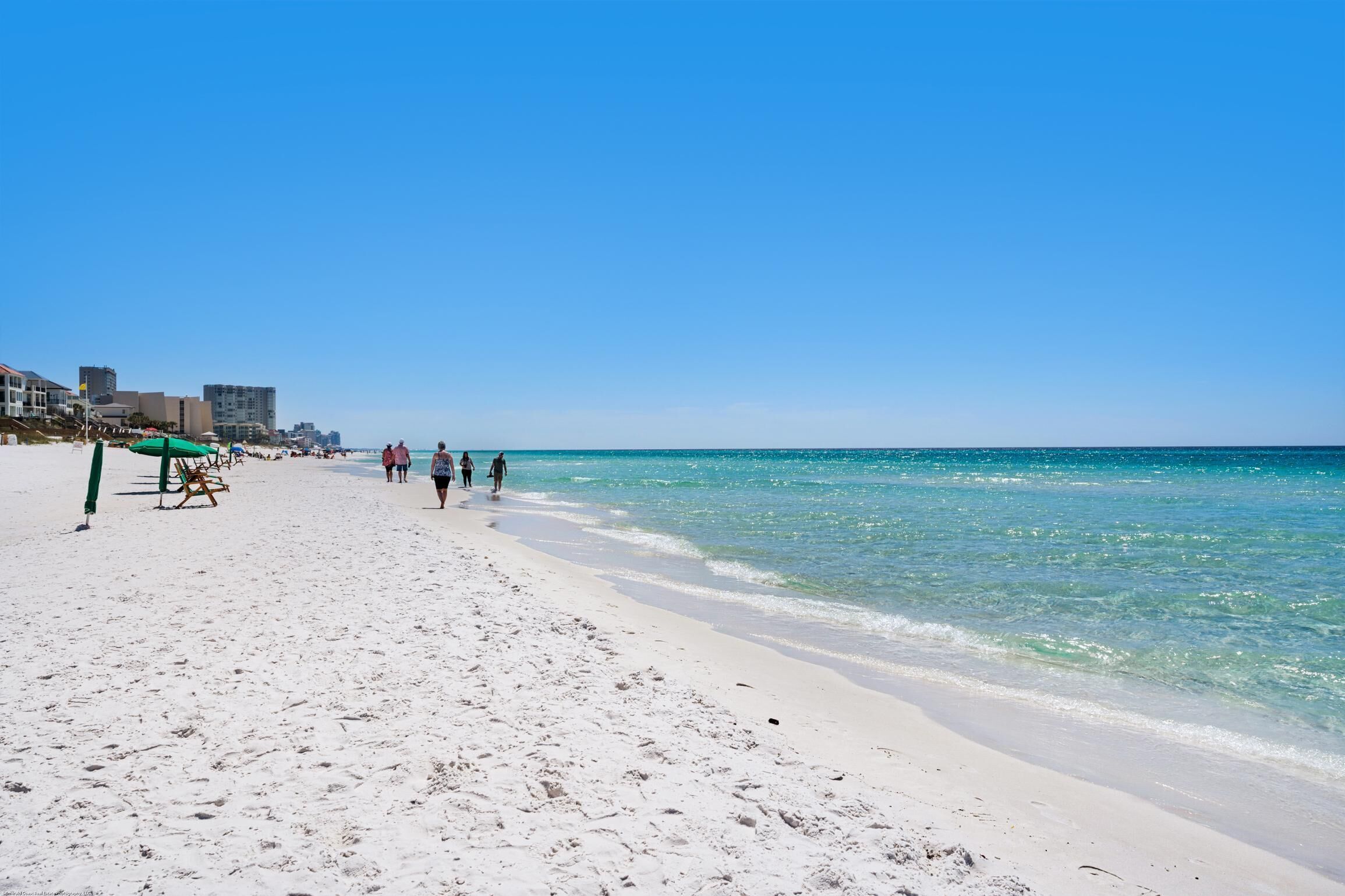 A beach with white sand, clear blue water, and a few people walking. There are buildings in the background.