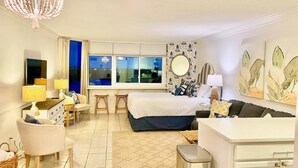 A hotel room with a bed, a desk, a chair, a small table, a trash bin, and a window with a view of the outside. - Top of the Gulf 808- Gulf Coast Getaway (Panama City Beach)