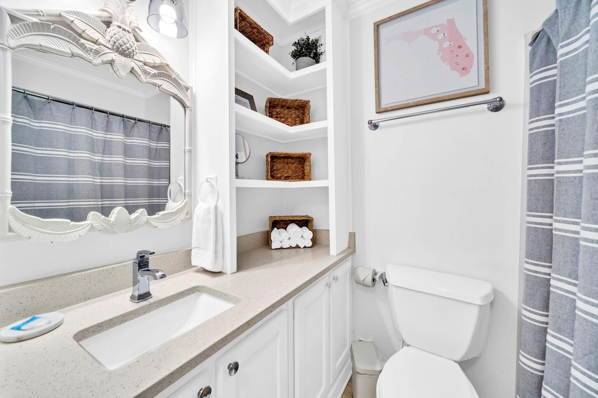 Un baño con un lavabo blanco, un inodoro y una cortina de ducha. Hay cestas de mimbre en estantes y un mapa enmarcado en la pared.