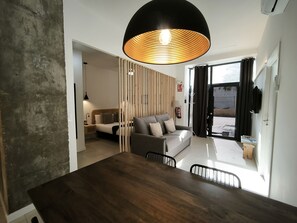 A modern hotel room with a wooden dining table, a sofa, and a large window with curtains.