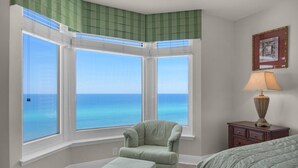 A room with a view of the ocean through large windows, a green armchair, a small ottoman, a nightstand with a lamp, and a framed picture on the wall. - Emerald Beach 1036 - Turquoise Turtle (Panama City Beach)