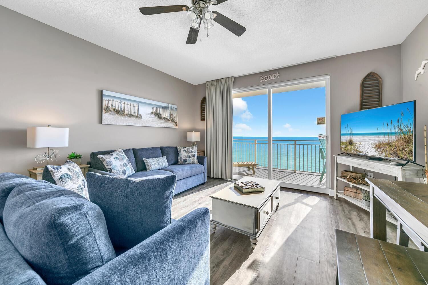 A modern living room with a sofa, a coffee table, and a TV. There is a balcony with a view of the ocean.