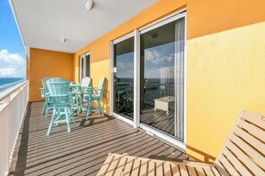 Un balcon avec des chaises turquoise et une table, une porte vitrée donnant sur une terrasse extérieure, et une vue sur l’océan.