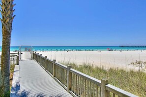 Une promenade en bois mène à une plage où l’eau est d’un bleu cristallin, et on aperçoit une jetée au loin.