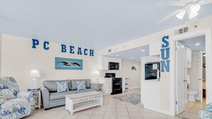 Un soggiorno con un divano grigio, una poltrona blu e un tavolino. C'è una televisione, una lampada e un forno a microonde. Sui muri si leggono le scritte 'PC BEACH' e 'SUN'.