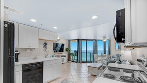 Una cucina moderna con mobili bianchi, lavastoviglie e piano cottura. C'è una zona pranzo con tavolo e sedie, e un'ampia finestra con vista sull'oceano.