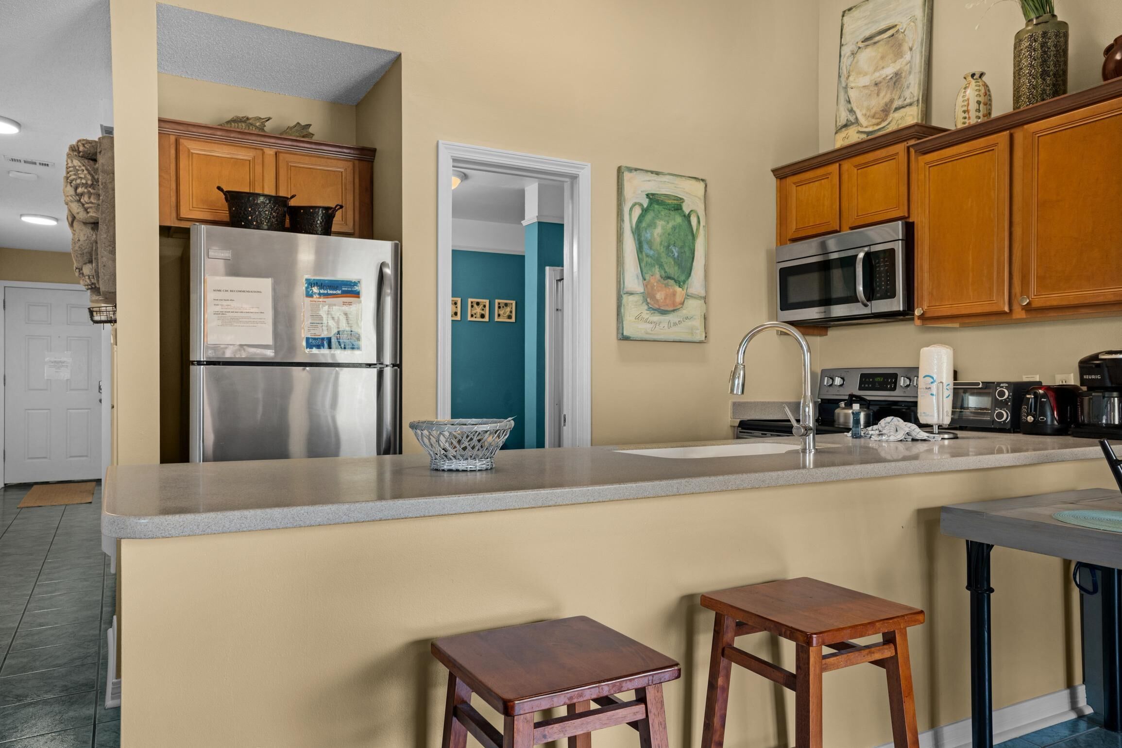 A kitchen with wooden cabinets, a stainless steel refrigerator, a microwave, and a sink. There are two wooden stools and a small table.