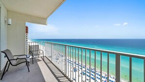 A balcony with two chairs, a railing, and a view of a beach with people and umbrellas.