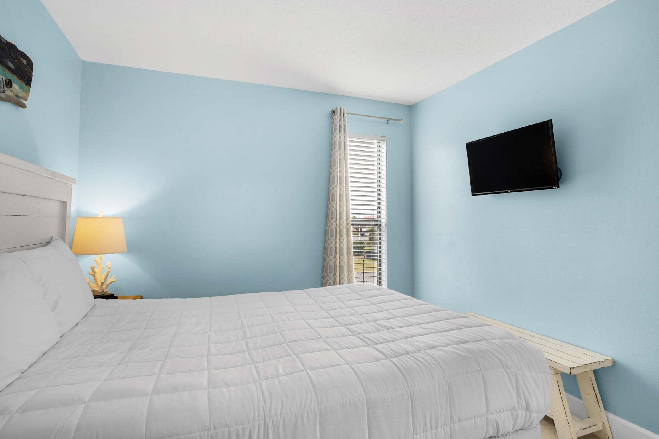 A bedroom with a bed, a bedside lamp, a TV mounted on the wall, and a window with blinds.
