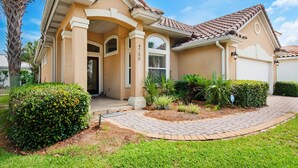 A two-story house with a beige facade, a tiled roof, and a well-maintained front yard with a brick pathway and various plants. - Wave N Sea (Destin)