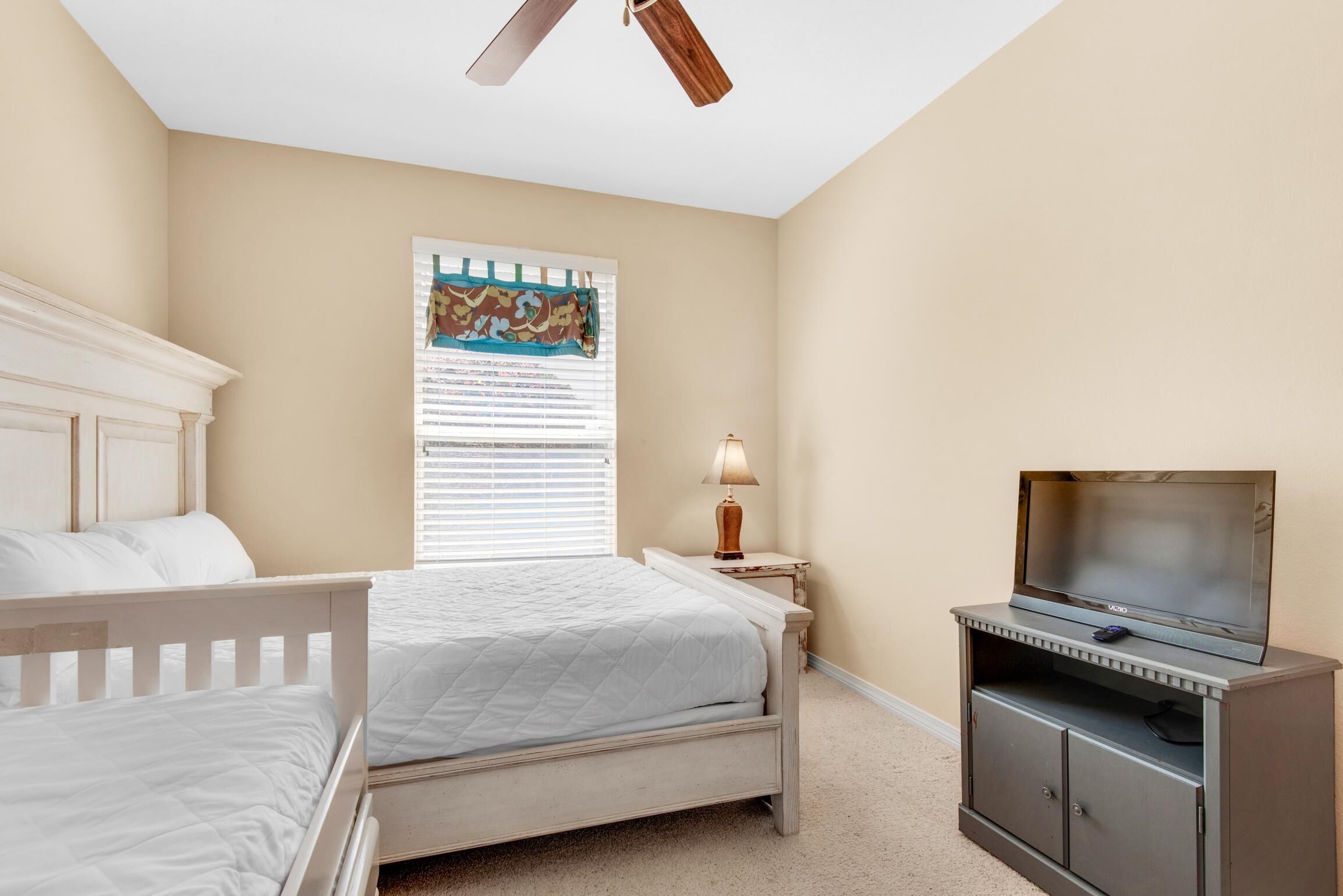 A bedroom with a bed, a TV on a stand, a ceiling fan, a window with blinds, and a wall-mounted lamp.