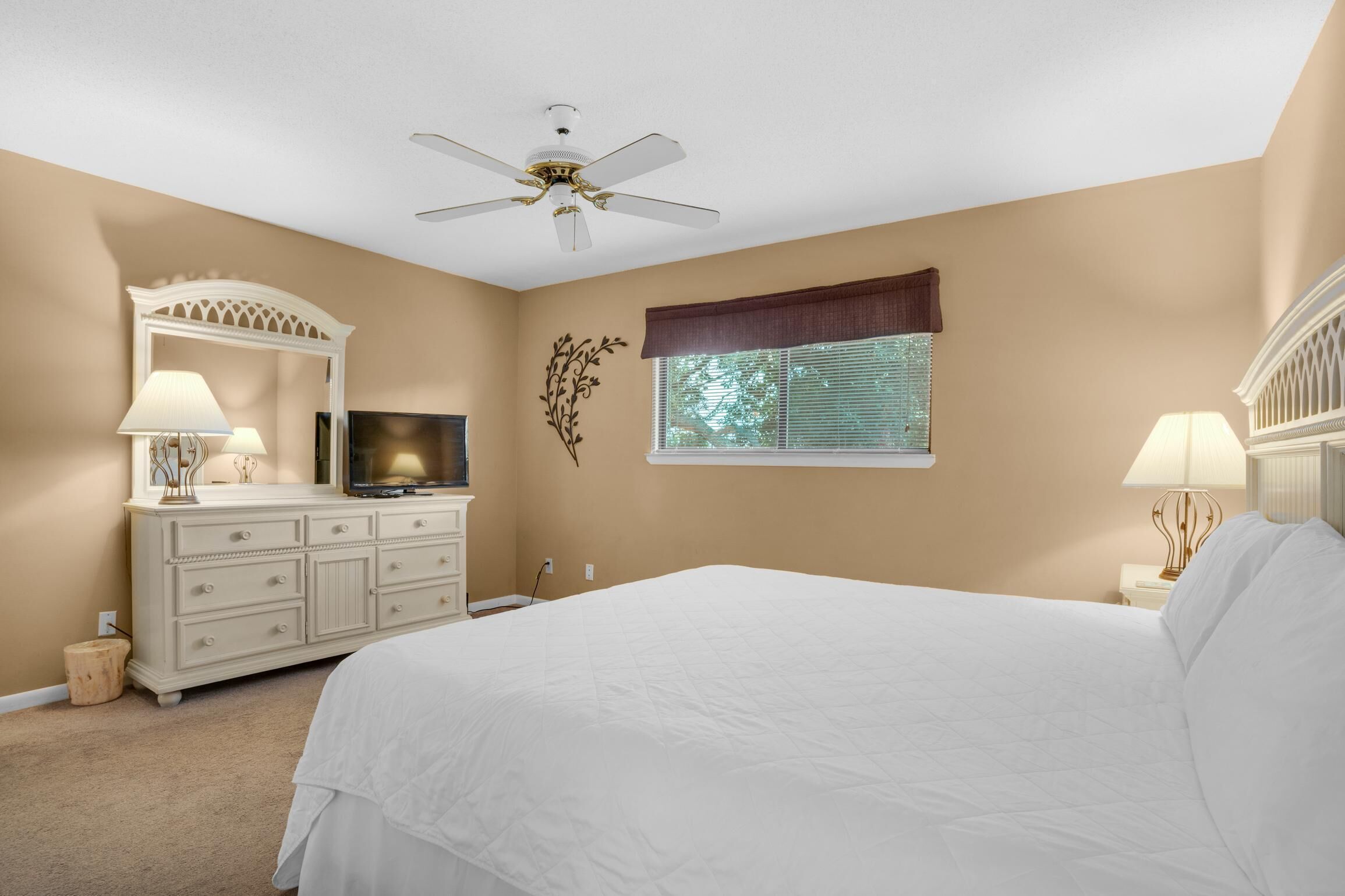 Ein Schlafzimmer mit Bett, Kommode, Fernseher, Deckenventilator, Fenster mit Jalousien und Wanddekoration.