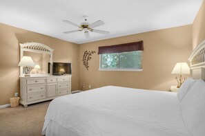 Une chambre à coucher comprenant un lit, une commode, un téléviseur, un ventilateur de plafond, une fenêtre avec des stores et des éléments de décoration murale.
