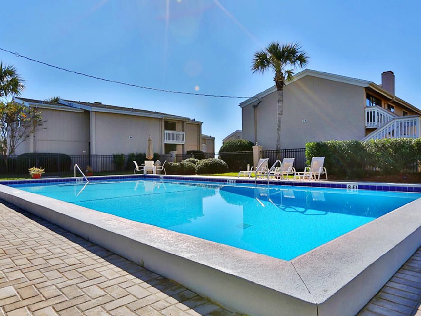 A rectangular swimming pool with clear blue water, surrounded by a tiled deck. There are lounge chairs and a palm tree nearby.