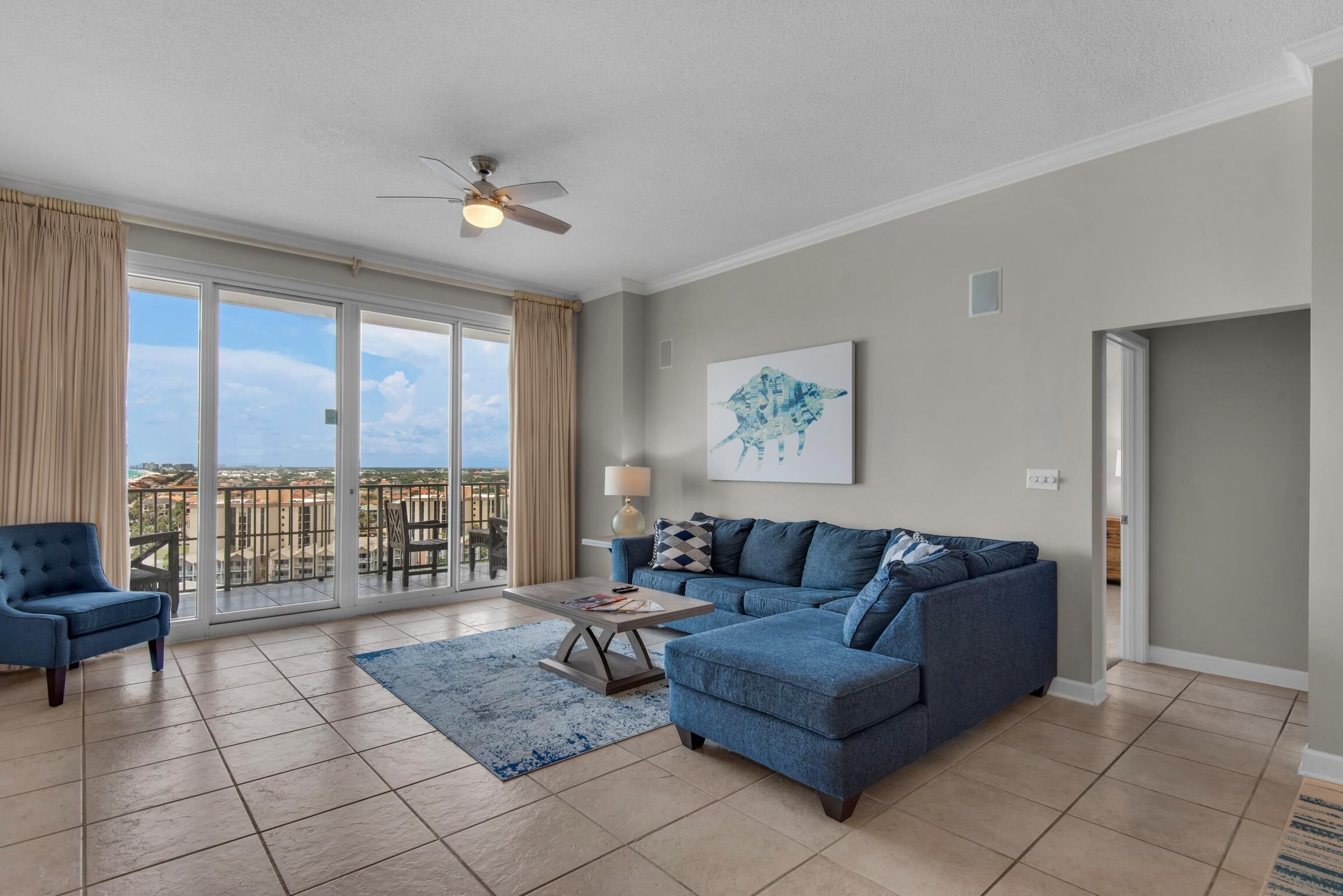 A spacious living room with a large sliding glass door leading to a balcony, a blue sofa set, a coffee table, and a ceiling fan.