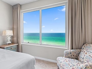 A hotel room with a large window offering a view of the ocean. There is a bed, a chair with a beach-themed cover, and a bedside table with a lamp. - Leeward Key 904 (Miramar Beach)