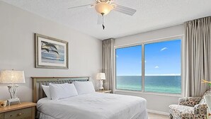 A hotel room with a large bed, a ceiling fan, a painting, a nightstand, a lamp, a chair, and a large window with a view of the ocean. - Leeward Key 904 (Miramar Beach)
