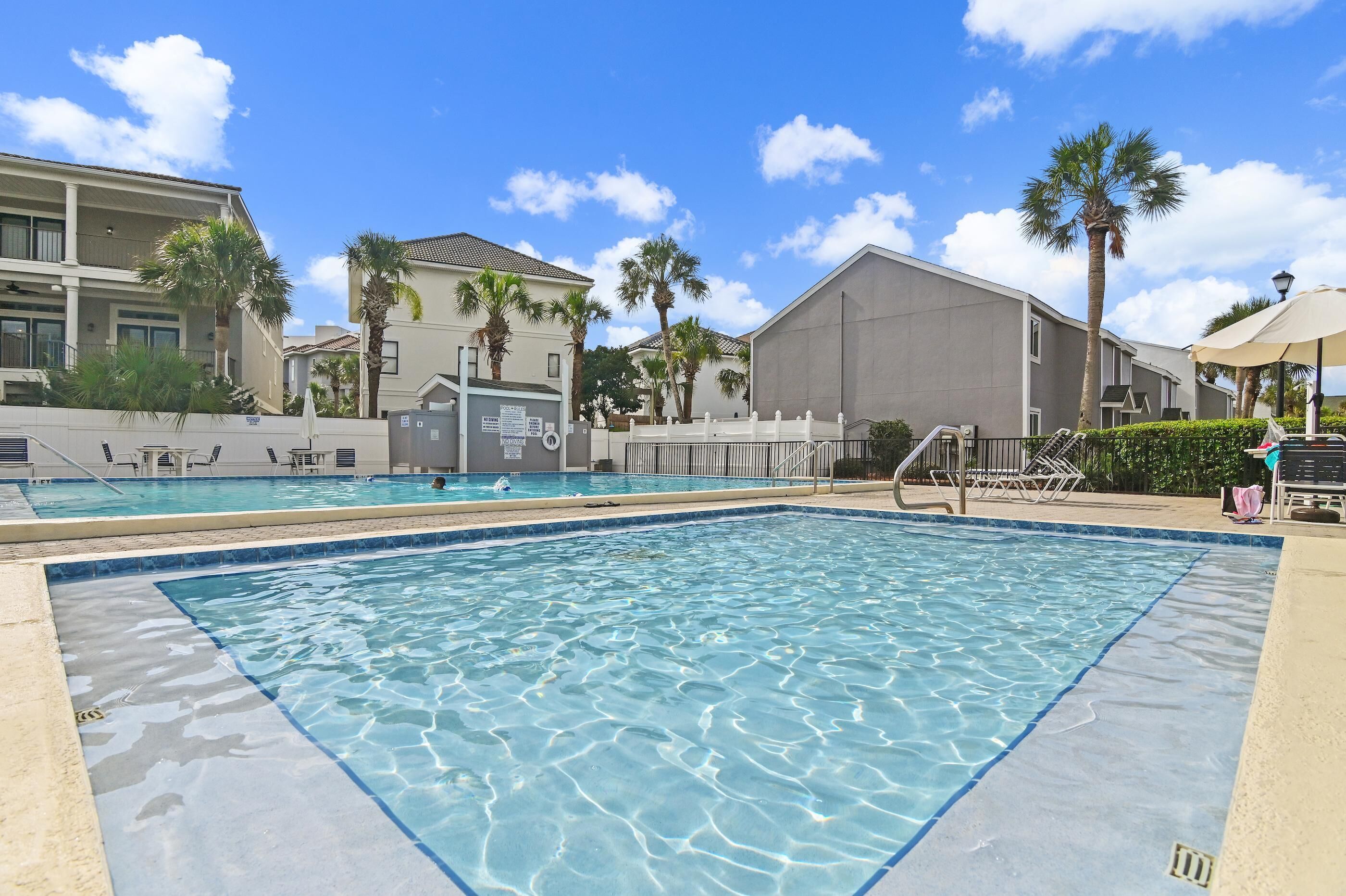 A swimming pool with clear water, surrounded by palm trees and a building with a balcony.