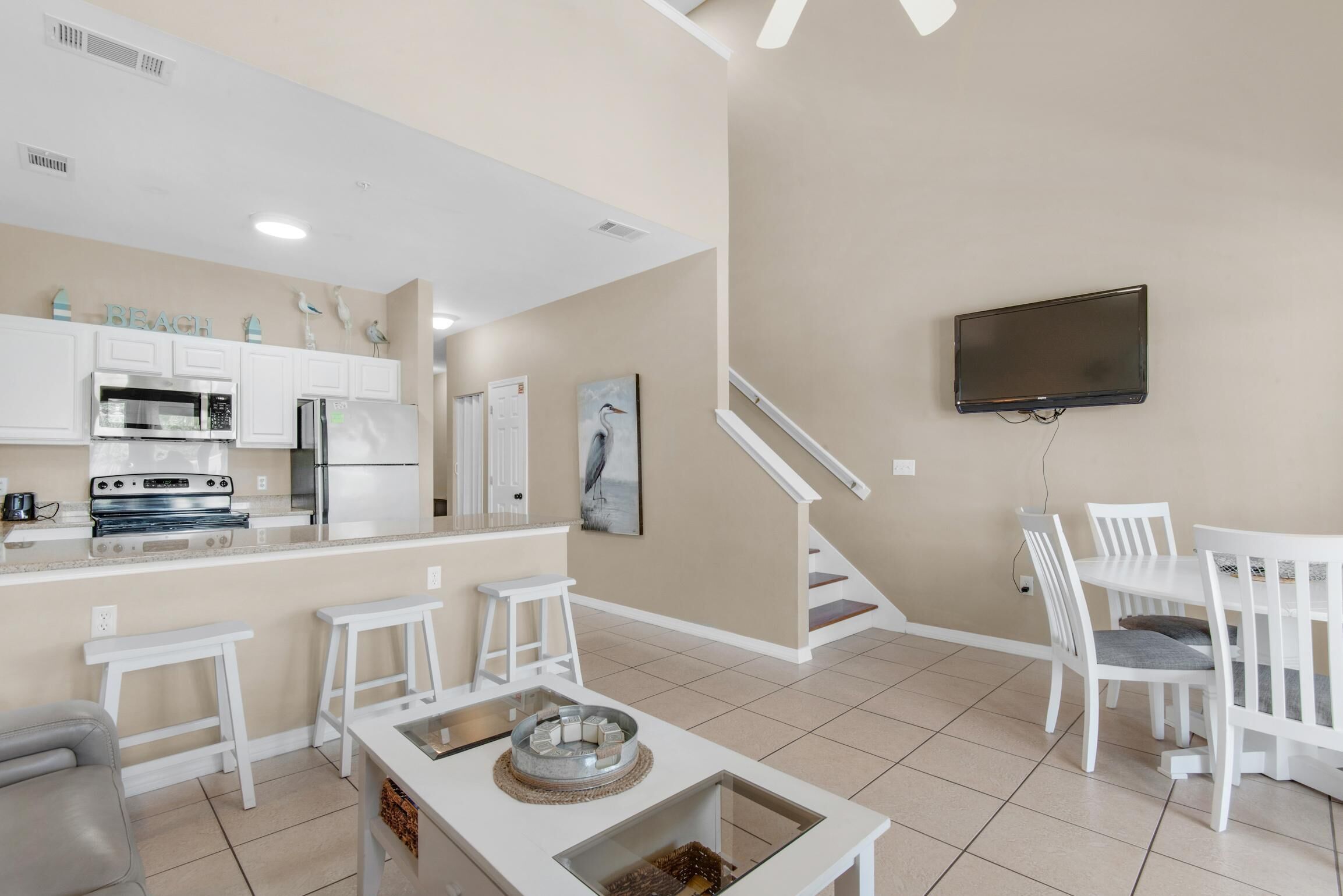 A modern kitchen with white cabinets, a built-in oven, and a refrigerator. There is a dining area with white chairs and a table, and a TV mounted on the wall.