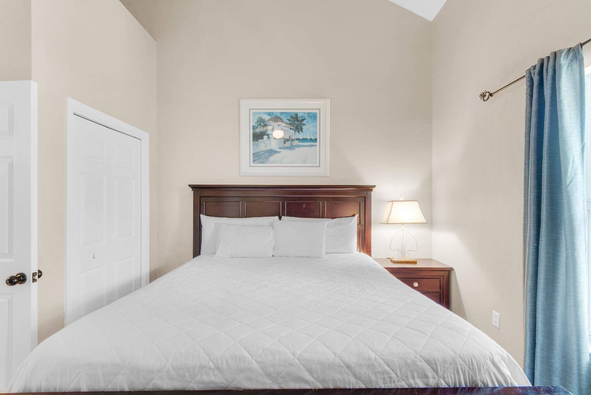 Un lit bien fait, recouvert d’une couette blanche, avec une tête de lit en bois, un tableau encadré au-dessus du lit, une table de chevet avec une lampe et un rideau bleu à la fenêtre.