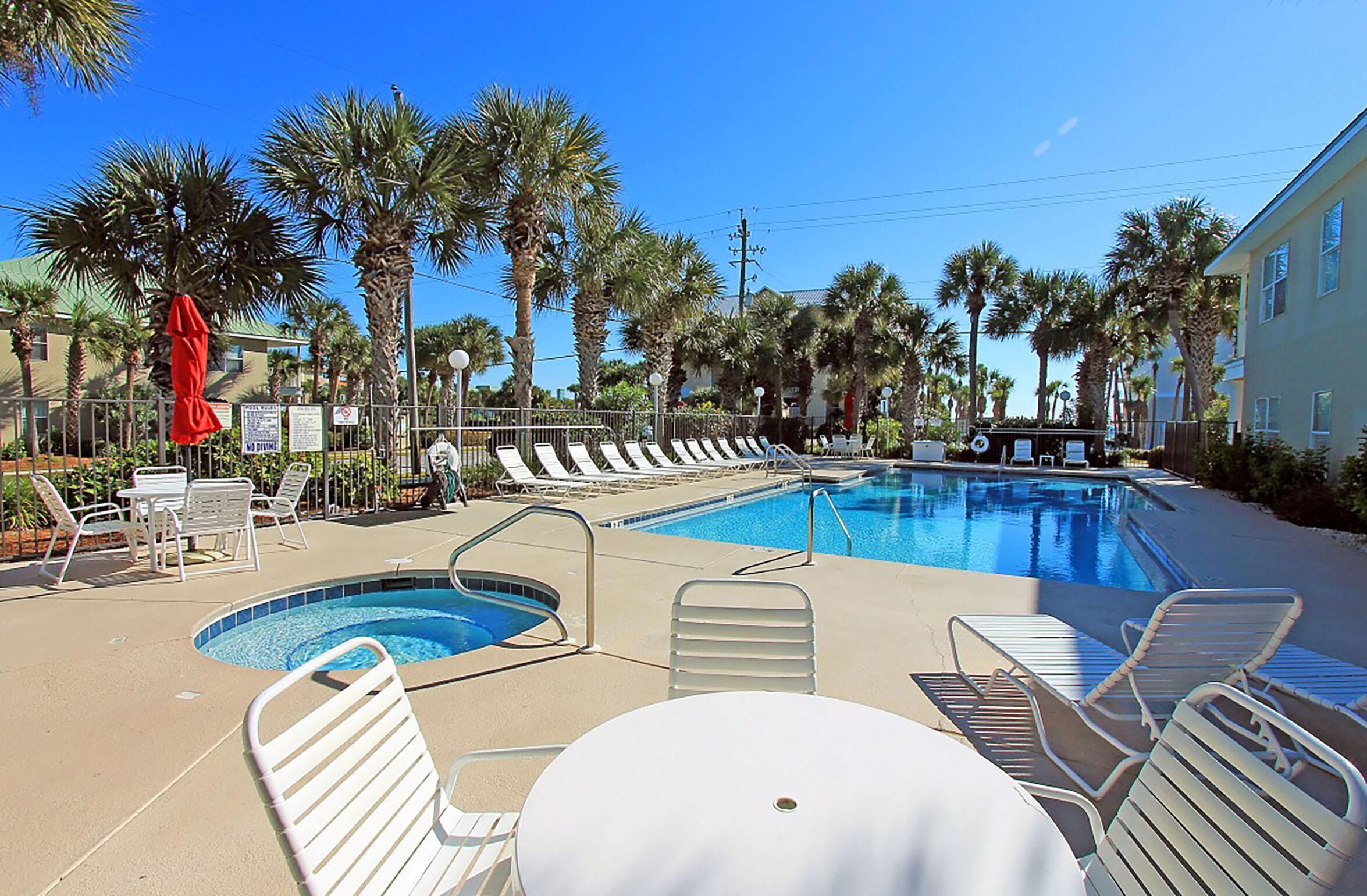 A pool area with lounge chairs and a small table, surrounded by palm trees and a building in the background.