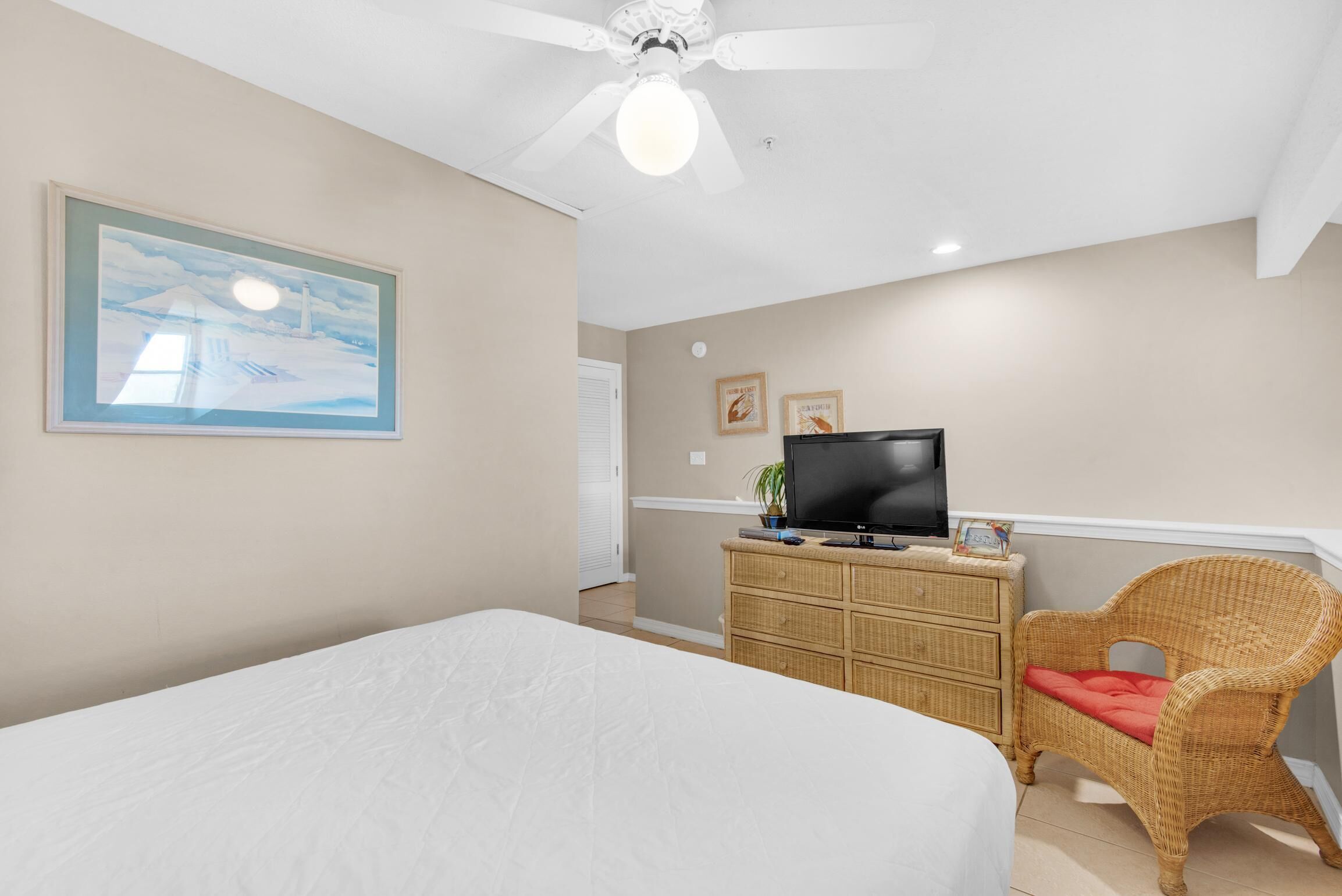 Un dormitorio con una cama, un televisor sobre una cómoda, una silla de mimbre y un ventilador de techo.