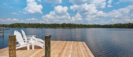 A wooden dock with white Adirondack chairs, a clear view of a lake, and a forested shoreline in the background.