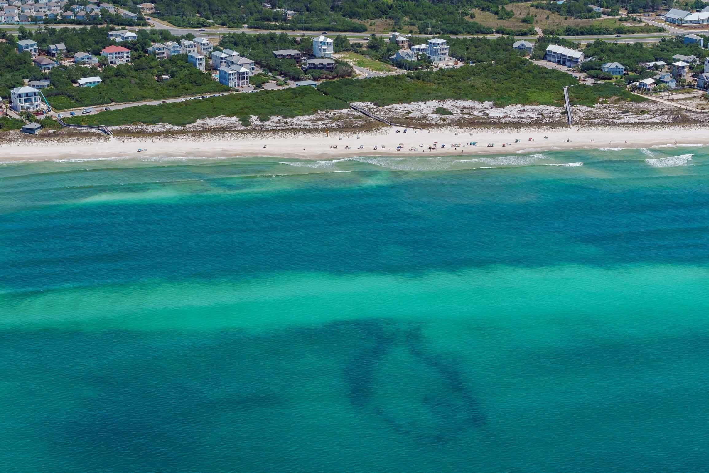 An aerial view of a coastal area with a beach, houses, and clear turquoise waters.