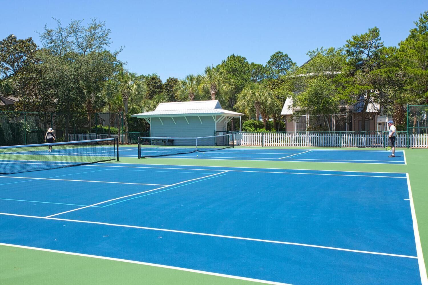 A tennis court with a net, surrounded by trees and a covered pavilion.