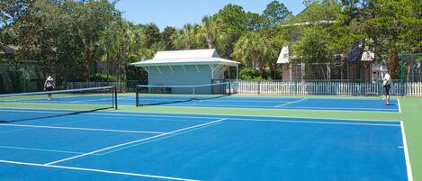 A tennis court with a net, surrounded by trees and a covered pavilion.