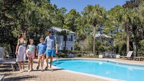 Uma piscina com um calçadão pavimentado, cercada por palmeiras e uma casa ao fundo.