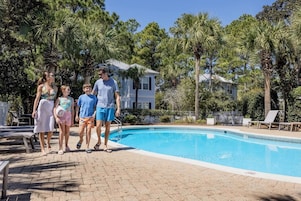 Uma piscina com um calçadão pavimentado, cercada por palmeiras e uma casa ao fundo.