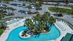 Une grande piscine bordée de palmiers et de chaises longues, entourée d’un parking et avec un bâtiment en arrière-plan.