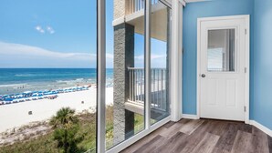 Une pièce dotée d’une grande porte-fenêtre coulissante qui donne sur un balcon surplombant une plage parsemée de parasols et fréquentée par des gens.