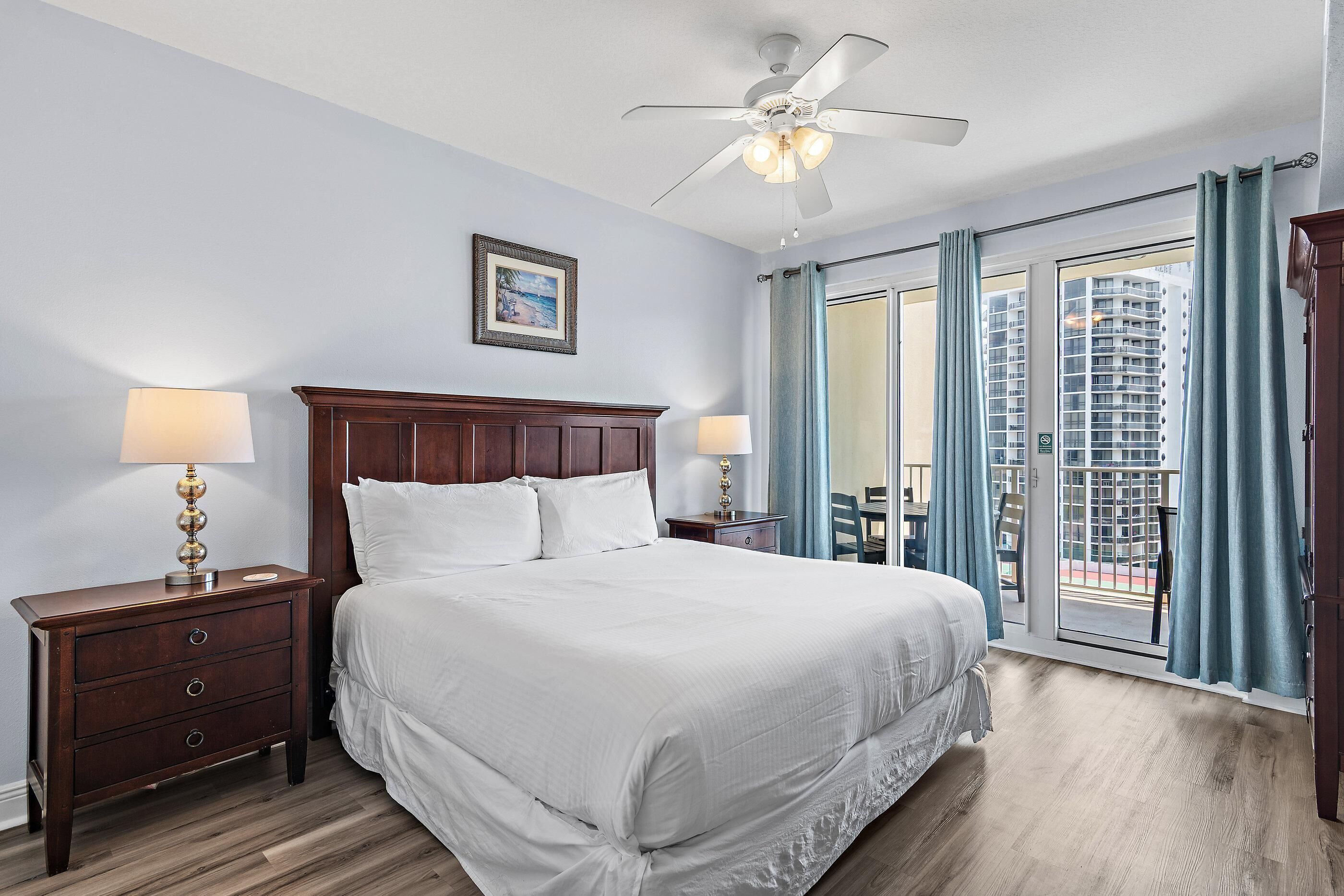 Habitación de hotel con una cama grande, cabecera de madera, dos mesitas de noche con lámparas, un ventilador de techo, un cuadro en la pared y vista a un edificio a través de la ventana.